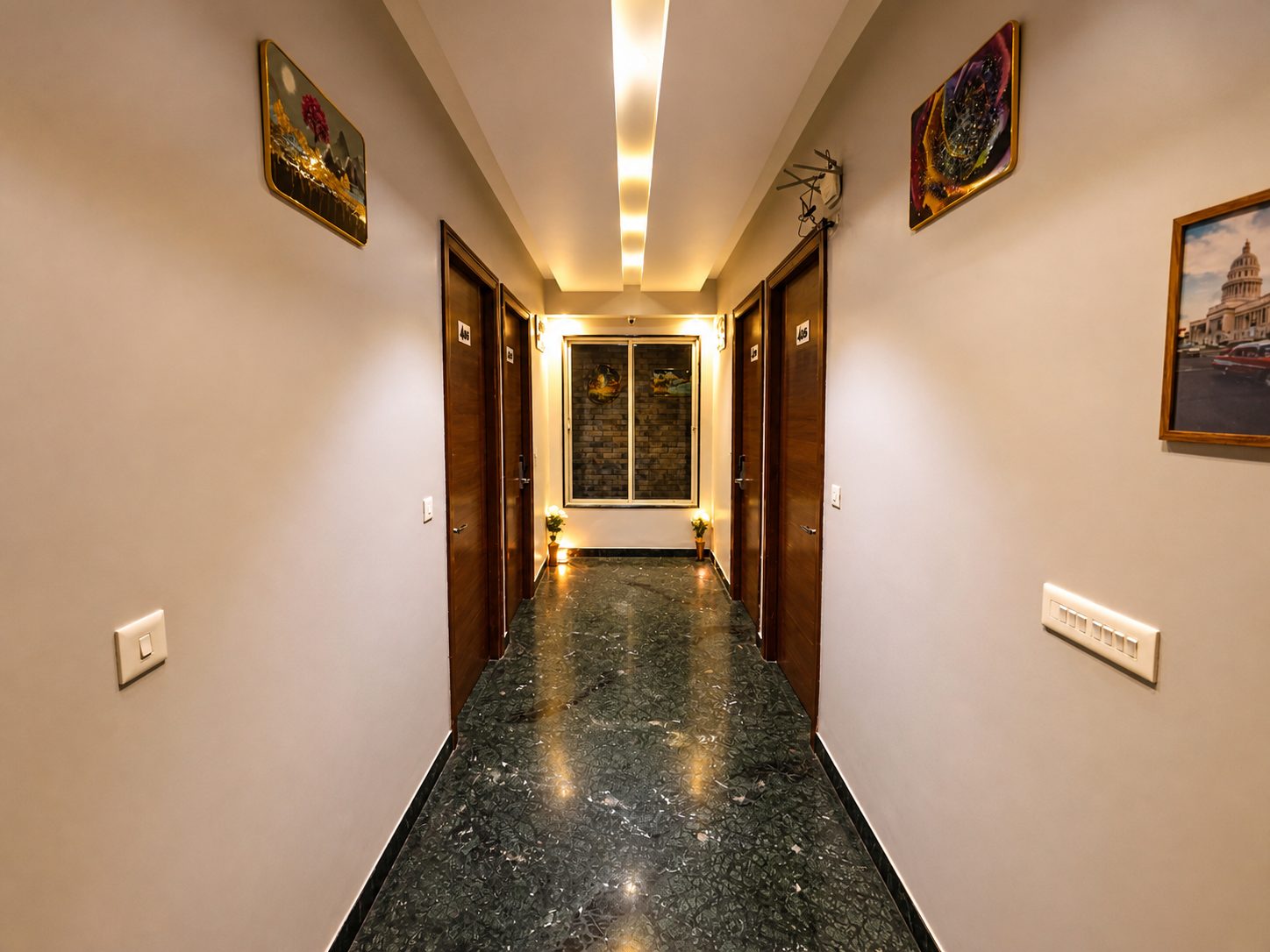 A guest corridor at night with four wooden doors, framed pictures on both walls, a strip of ceiling light and a window at the far end.