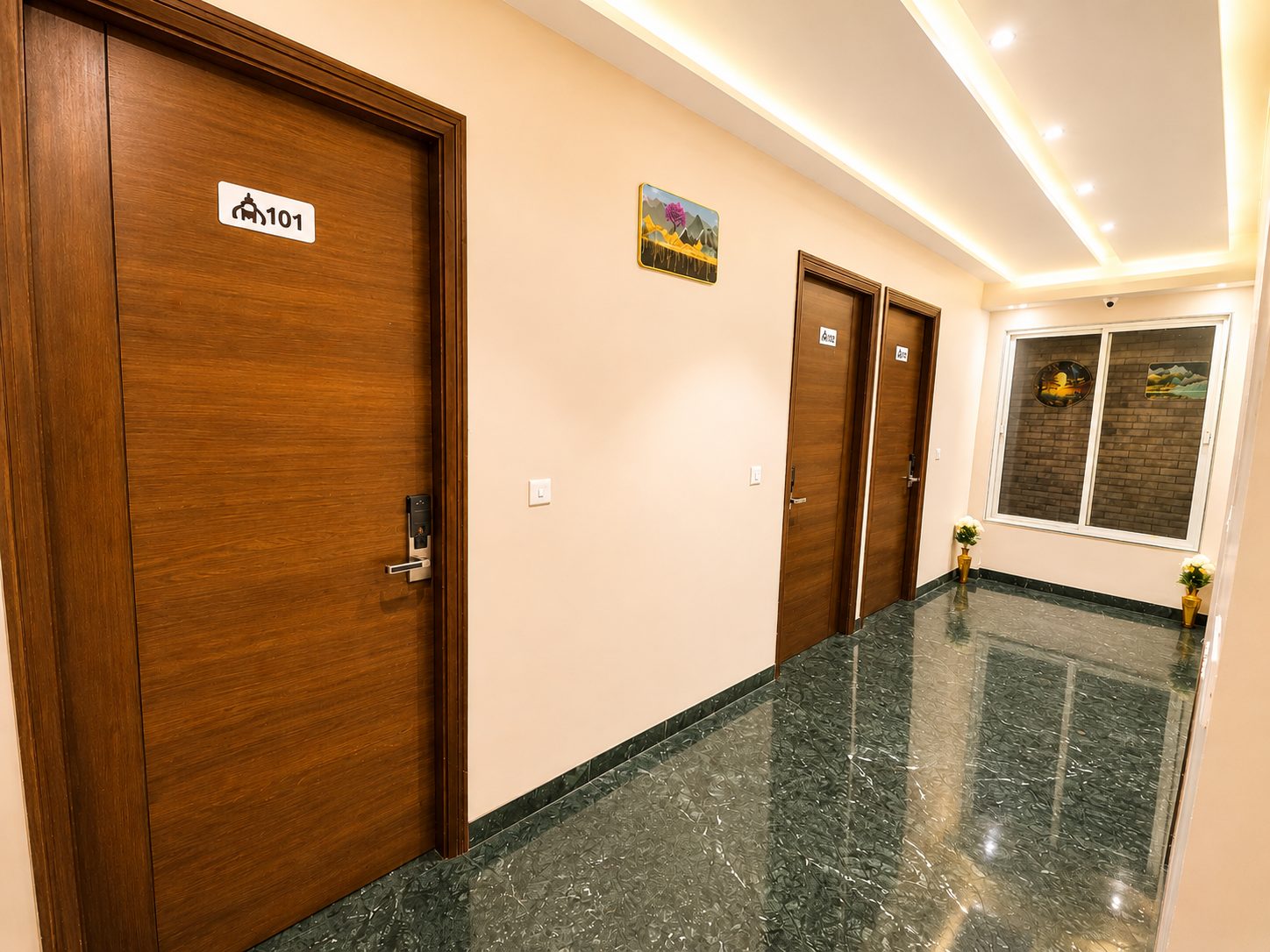 A guest corridor on green marble with wooden doors, room 101 in front, a painting on the wall and a window at the end.
