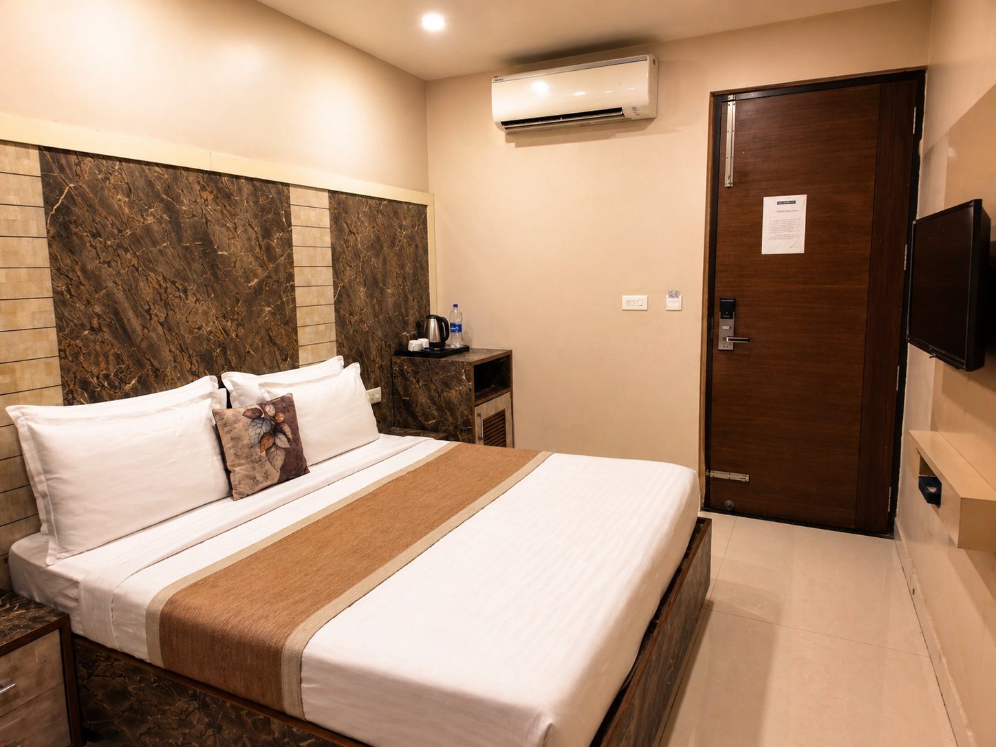 A guest room with a double bed against a brown marble-pattern wall, a kettle and water on the side table, an air conditioner above and a television opposite.