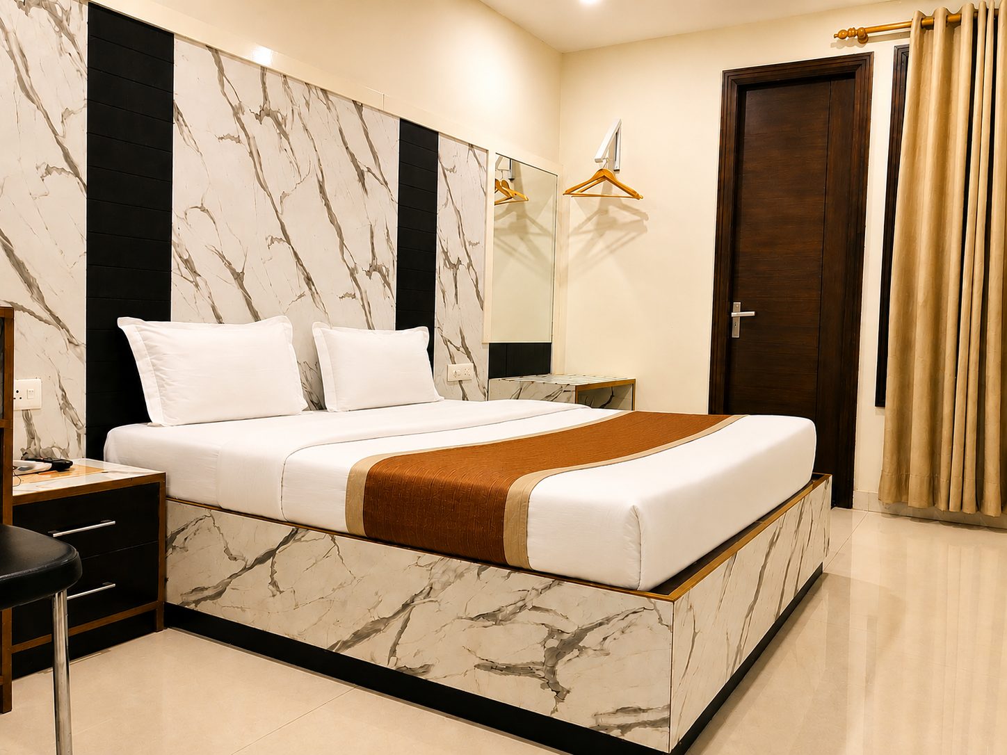 A guest room with a double bed set in white marble-pattern panelling with black strips, a rust-brown runner, hangers by a mirror and gold curtains.